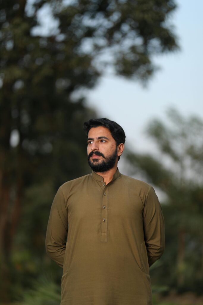 A thoughtful bearded man in a green shirt stands outdoors in Kasur, Pakistan.