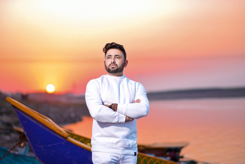 A man in white casual attire standing at sunset by a colorful boat and calm sea.