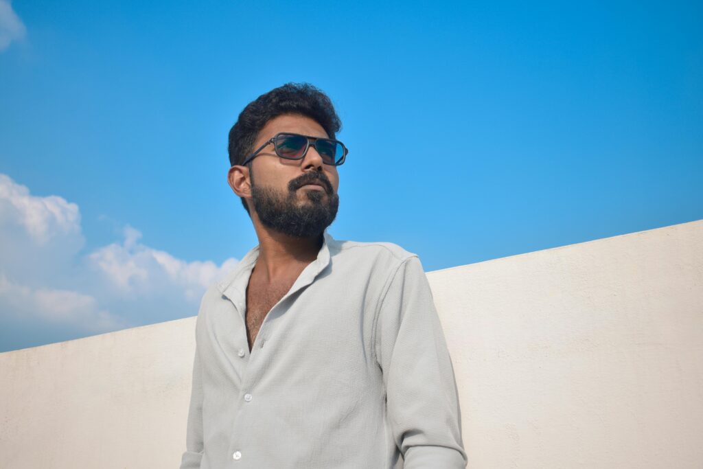 Confident man wearing sunglasses, posing outdoors against a bright blue sky.